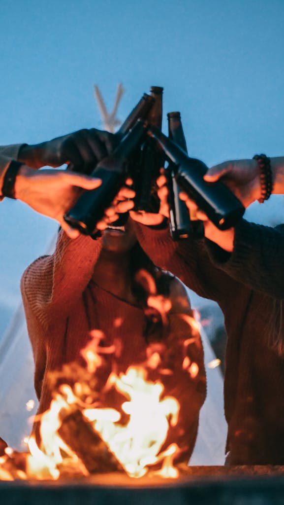 A group of friends enjoys a bonfire, toasting beer bottles, and celebrating togetherness.