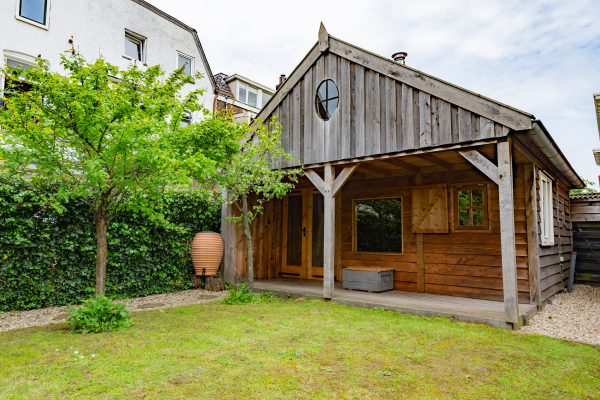 Modern barn-style cottage with weathered wood exterior, covered porch, and lush green garden landscaping.